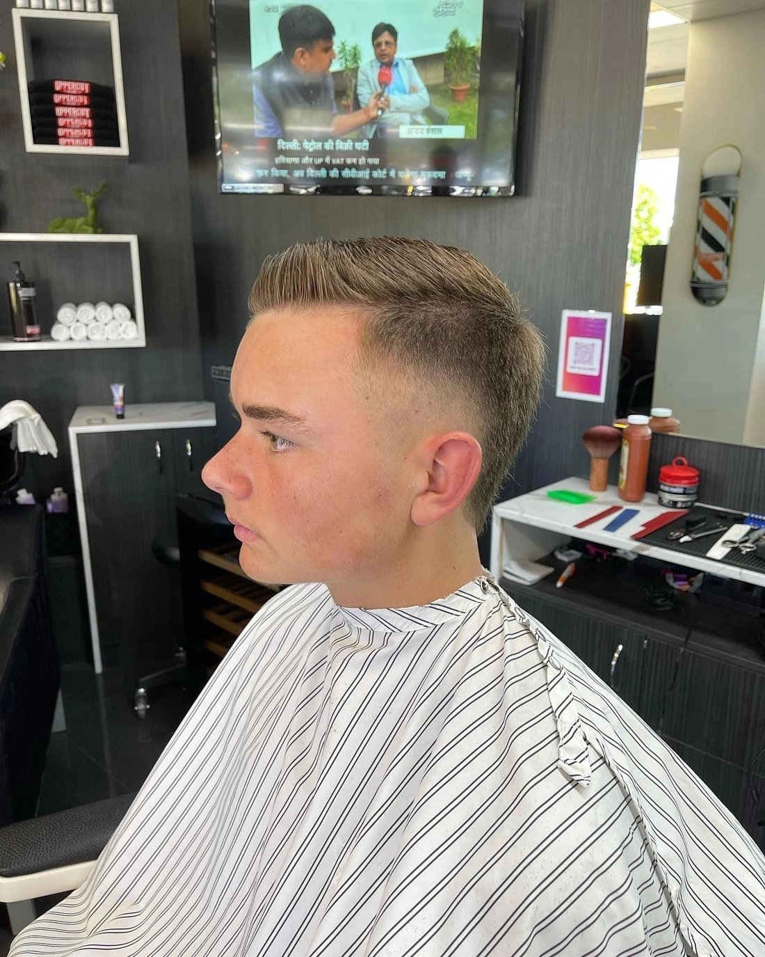 A client in the chair with a textured top and a taper fade, the barber pole and product shelves behind him.