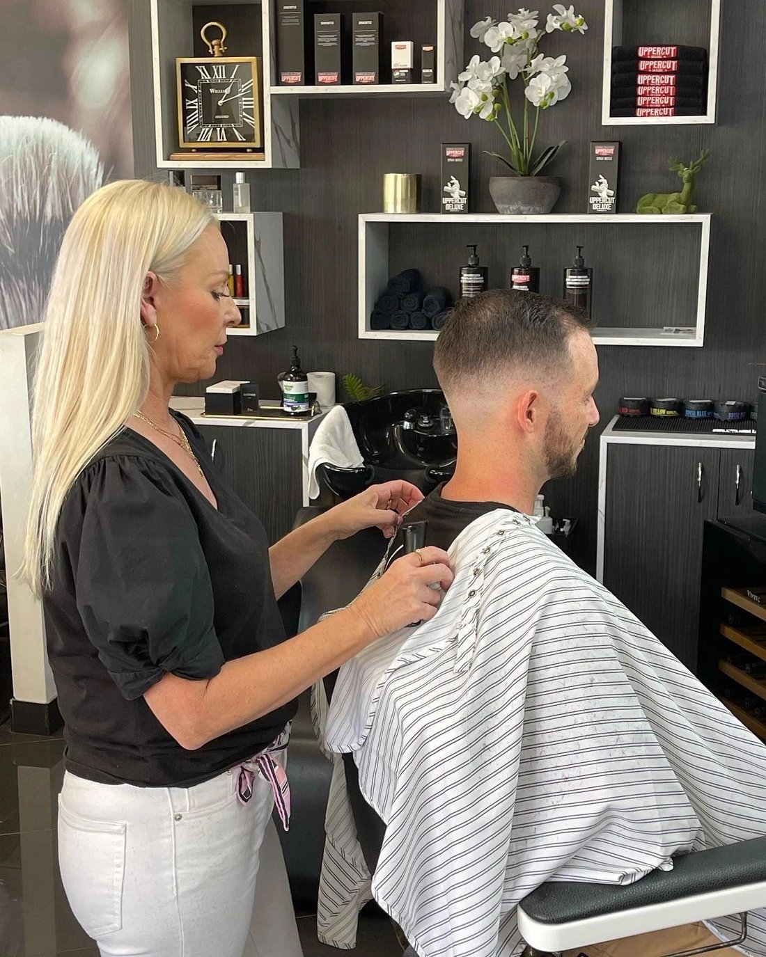A barber fastening the cape on a client at the chair, with the product shelves and wash basin behind.